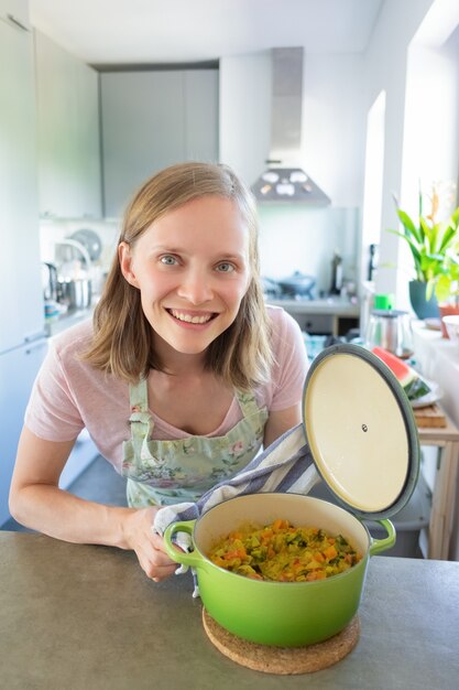 Mulher jovem feliz desfrutando de cozinhar na cozinha dela, abrindo a panela com farinha de vegetais, encostado na mesa e sorrindo para a câmera. Tiro vertical. Conceito de blogueiro de comida