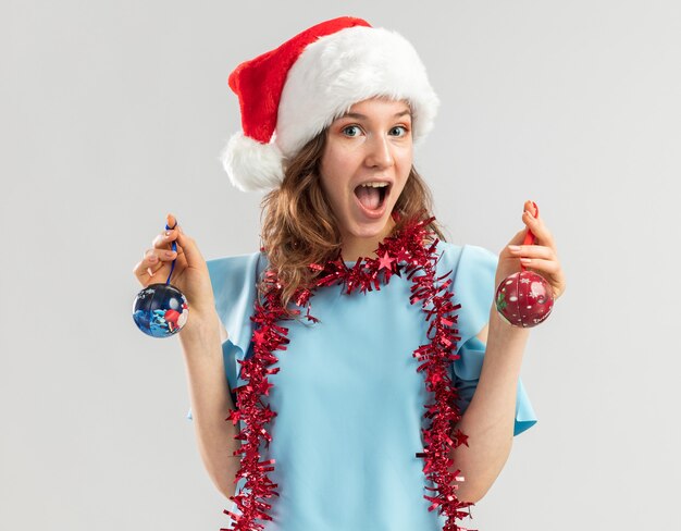 Mulher jovem feliz e animada com uma blusa azul e um chapéu de Papai Noel com enfeites em volta do pescoço segurando bolas de Natal e sorrindo alegremente