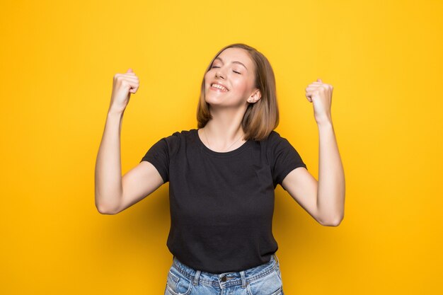 Mulher jovem feliz e bem-sucedida com as mãos levantadas, gritando e comemorando o sucesso na parede amarela
