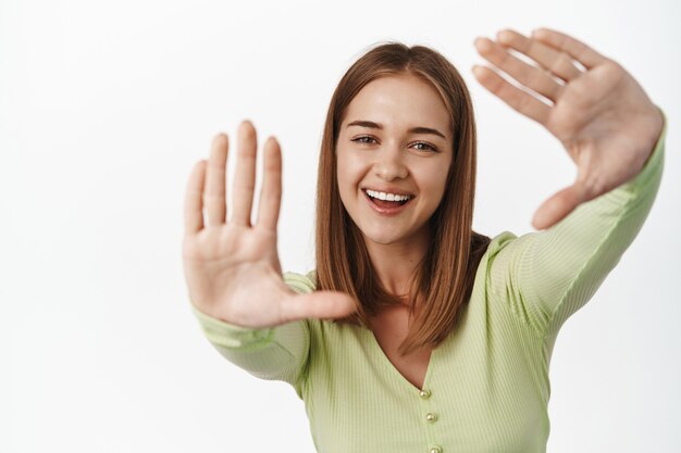 Mulher jovem feliz e brilhante rindo, sorrindo e olhando através de molduras de mão, imaginando algo