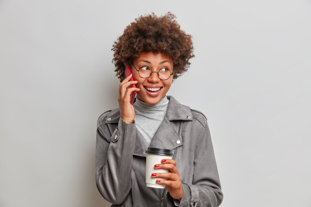 Mulher jovem feliz e despreocupada com cabelo afro, conversa animada ao telefone, desvia o olhar, segura uma xícara descartável de café, usa óculos redondos e jaqueta cinza