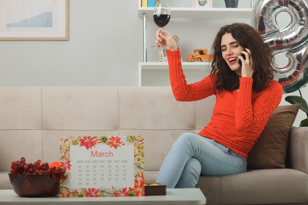 Foto grátis mulher jovem feliz e satisfeita em roupas casuais, sorrindo alegremente sentada em um sofá com uma taça de vinho falando no celular na sala de estar iluminada, celebrando o dia internacional da mulher, 8 de março