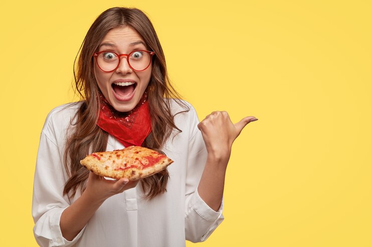 Foto grátis mulher jovem feliz e satisfeita olha com felicidade, aponta com o polegar de lado para o espaço livre, come pizza, mostra a direção, fica de queixo caído, exclama de felicidade, isolada sobre parede amarela