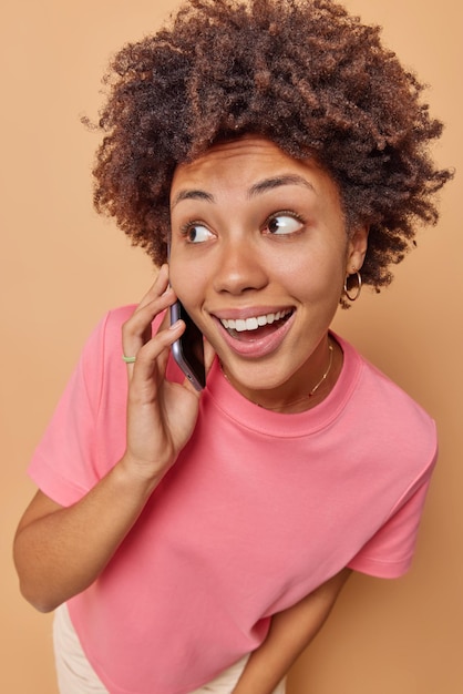 Mulher jovem feliz e surpresa conversando ao telefone com uma expressão de alegria que passa o tempo livre fofocando com um amigo via celular usa uma camiseta rosa isolada sobre um fundo bege