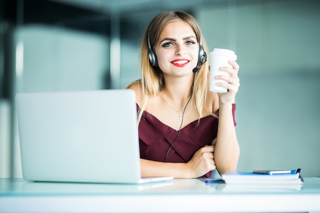 Mulher jovem feliz em fones de ouvido em call center e bebendo café no escritório.