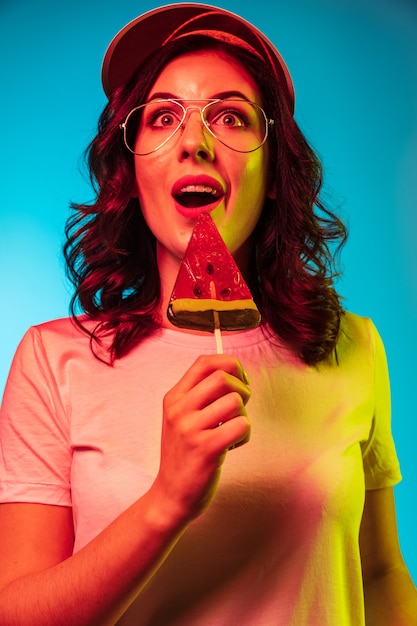 Foto grátis mulher jovem feliz em óculos de sol comendo doces e sorrindo sobre néon azul da moda