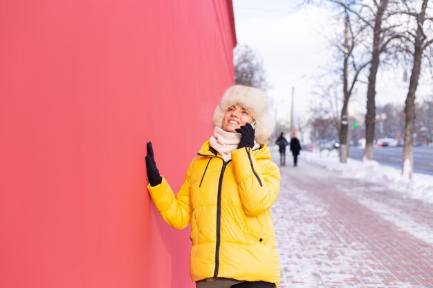 Mulher jovem feliz em um fundo de uma parede vermelha com roupas quentes em um dia ensolarado de inverno, sorrindo e falando ao telefone em uma calçada de uma cidade nevada