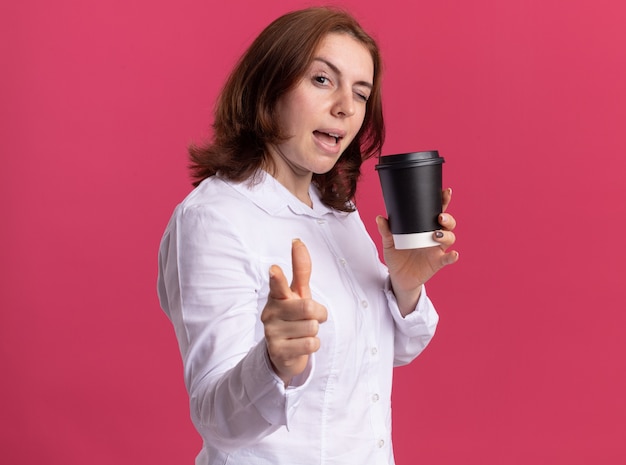 Mulher jovem feliz em uma camisa branca segurando uma xícara de café apontando com o dedo indicador na frente, sorrindo e piscando em pé sobre a parede rosa