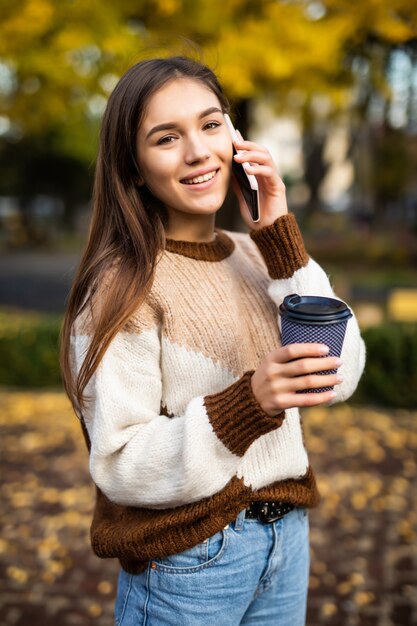 Mulher jovem feliz falando ao telefone, segurando uma xícara de café para viagem e sorrindo.