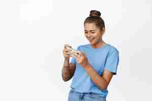 Foto grátis mulher jovem feliz jogando videogame móvel e sorrindo, vestindo camiseta azul, em pé contra um fundo branco