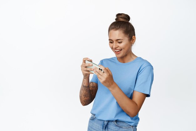 Mulher jovem feliz jogando videogame móvel e sorrindo, vestindo camiseta azul, em pé contra um fundo branco