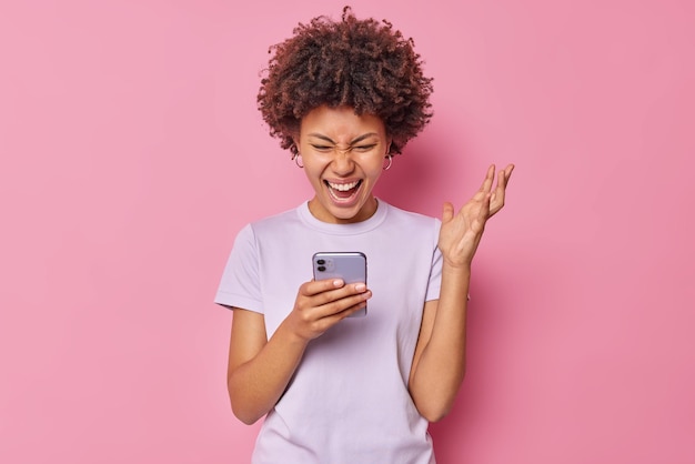 Mulher jovem feliz mantém a mão levantada e reage a algo positivo concentrado na tela do smartphone segura o celular vestido com uma camiseta casual isolada sobre a parede rosa