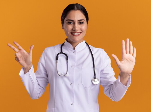 Mulher jovem feliz, médica com jaleco com estetoscópio no pescoço, olhando para a frente com um grande sorriso no rosto, mostrando o número oito em pé sobre uma parede laranja