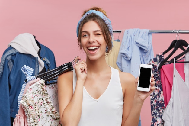 Foto grátis mulher jovem feliz, mostrando a tela em branco copyspace do telefone móvel, segurando cabides de elegantes peças de roupa e piscando