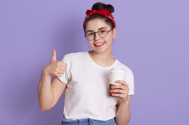 Mulher jovem feliz mostrando o polegar e segurando o café para viagem, olhando sorrindo diretamente para a câmera, usando traje casual e faixa vermelha