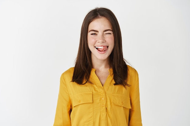 Mulher jovem feliz mostrando uma língua boba e piscando para a câmera, expressando positividade e alegria, em uma blusa amarela na parede branca