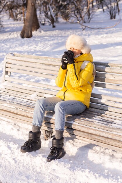 Mulher jovem feliz no inverno com roupas quentes em um parque nevado em um dia ensolarado se senta nos bancos e aproveita o ar fresco e o café sozinha