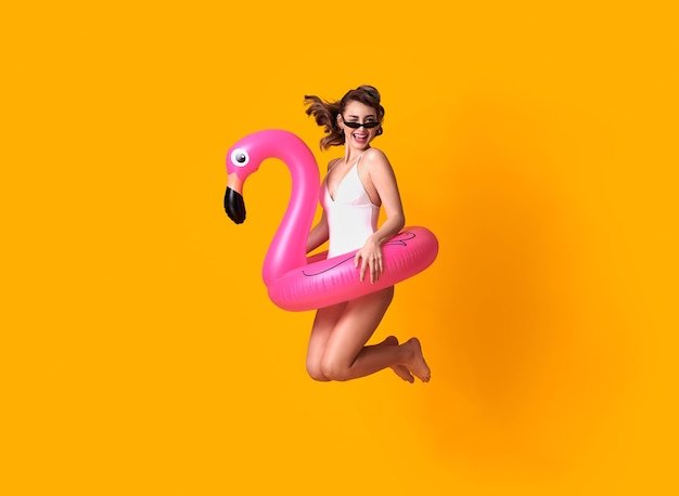 Mulher jovem feliz pulando na parede amarela, vestida em trajes de banho, segurando a praia de anel de borracha de flamingo.