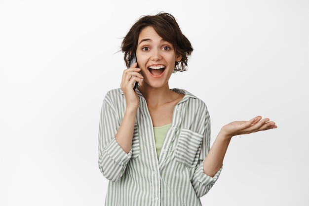 Mulher jovem feliz recebe telefonema, falando no celular e sorrindo surpreso, de pé sobre fundo branco.