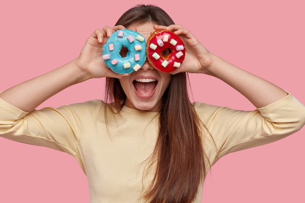 Foto grátis mulher jovem feliz segura dois donuts saborosos contra os olhos, abre a boca de espanto, gosta de sobremesa doce, isolada sobre o espaço rosa