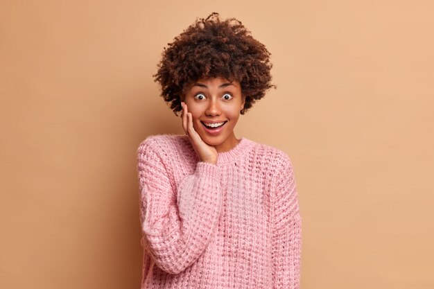 Mulher jovem feliz surpresa com cabelo afro parece animado e sorriso largo mantém a mão no rosto vestida em poses casuais de macacão de malha contra a parede bege do estúdio