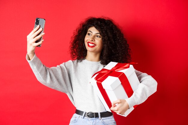 Mulher jovem feliz tomando selfie com seu presente de dia dos namorados, segurando um presente e fotografando em smartphone, posando em fundo vermelho.