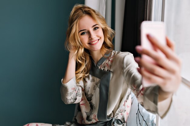 Mulher jovem feliz vestindo pijama de seda tomando selfie pela manhã, sentado no parapeito da janela. Ela tem cabelos loiros ondulados. Sala com parede turquesa.