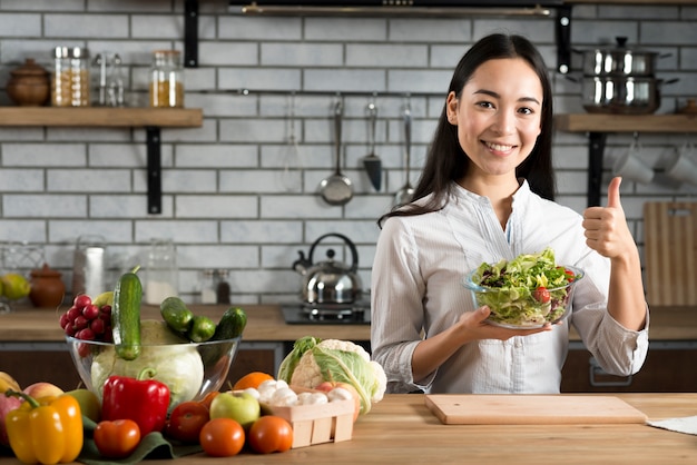 Mulher jovem, ficar, perto, contador cozinha, mostrando, polegar cima, sinal, segurando, legumes, salada