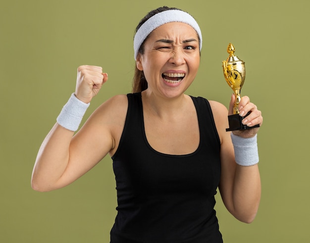 Mulher jovem fitness com bandana e braçadeiras segurando um troféu com raiva e frustrada cerrando os punhos