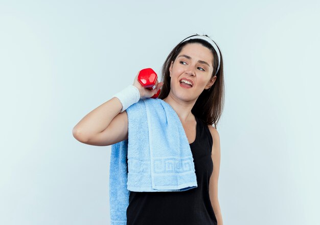 Mulher jovem fitness com bandana e toalha no ombro, malhando com halteres, parecendo confiante em pé sobre uma parede branca