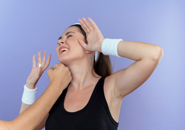 Mulher jovem fitness com bandana levando um soco na cara, de pé sobre um fundo azul