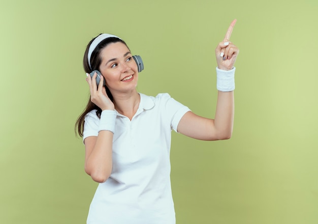 Mulher jovem fitness com bandana olhando para o lado apontando com o dedo para o lado sorrindo em pé sobre um fundo claro