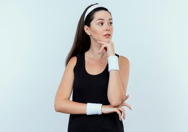 Mulher jovem fitness com bandana olhando para o lado com a mão no queixo pensando em pé sobre um fundo branco