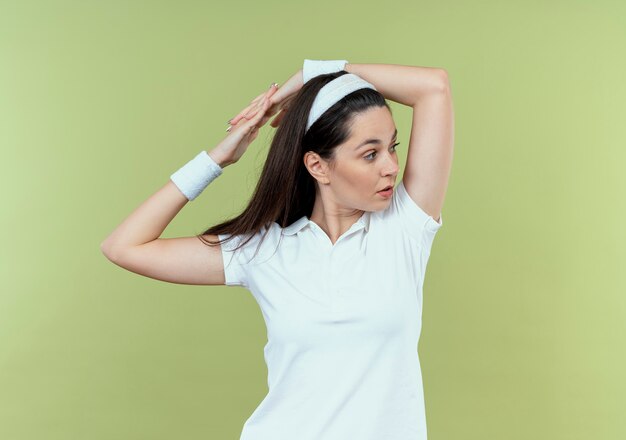 Mulher jovem fitness com bandana olhando para o lado, esticando as mãos em pé sobre um fundo claro