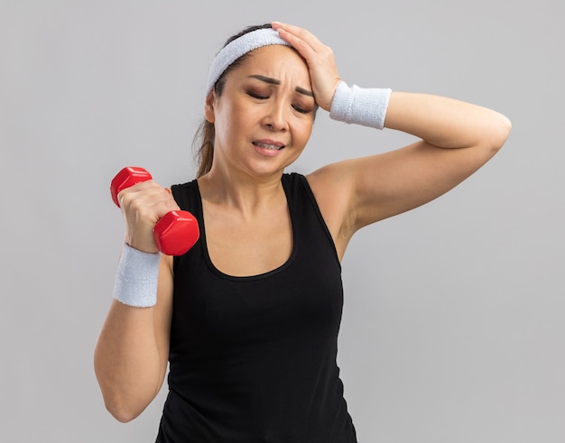 Mulher jovem fitness com bandana segurando halteres, fazendo exercícios, parecendo confusa com a mão na cabeça por engano