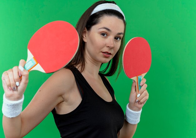 Mulher jovem fitness com bandana segurando raquetes de tênis de mesa, olhando para a câmera com expressão confiante em pé sobre fundo verde