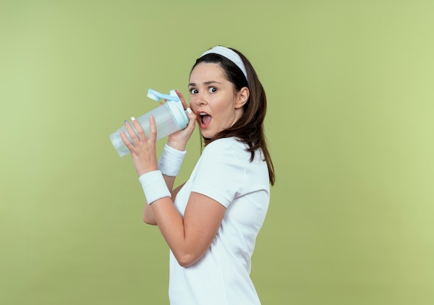 mulher jovem fitness com bandana segurando uma garrafa de água e surpresa em pé sobre a parede de luz