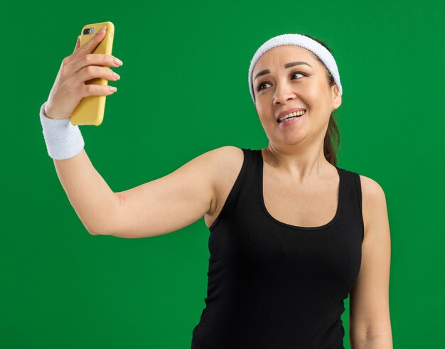 Mulher jovem fitness com faixa para a cabeça e braçadeiras, feliz e positiva, sorrindo fazendo selfie usando smartphone em pé sobre a parede verde