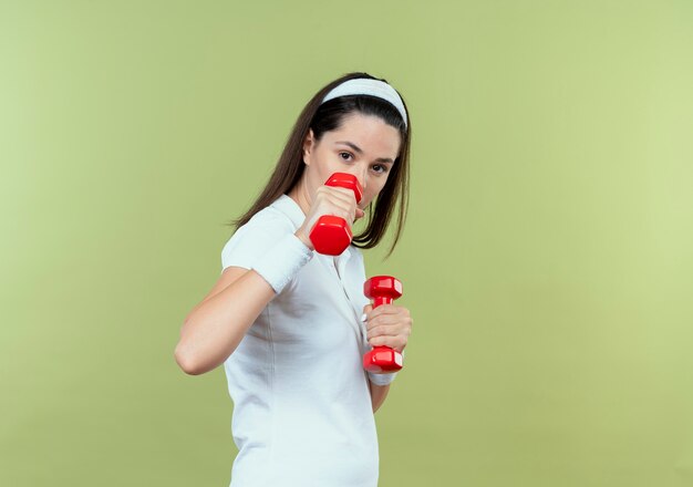 Mulher jovem fitness com faixa para a cabeça e malhando com halteres e expressão confiante em pé sobre a parede de luz