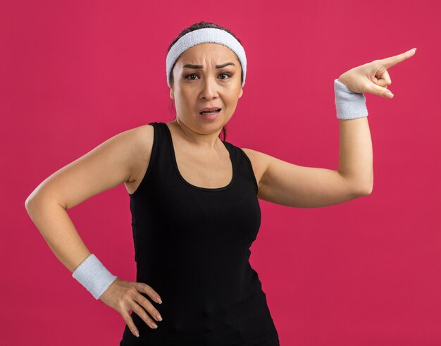Mulher jovem fitness com fita para a cabeça confusa, apontando com o dedo indicador para o lado em pé sobre a parede rosa