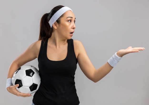 Mulher jovem fitness com fita para a cabeça e braçadeiras segurando uma bola de futebol, olhando de lado, confusa com o braço estendido em pé sobre uma parede branca