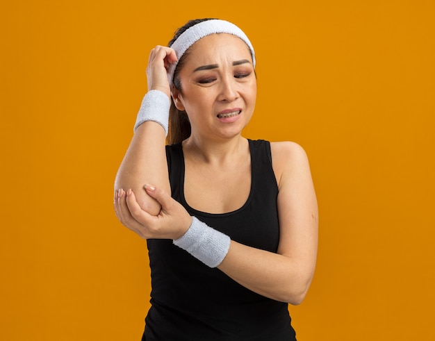 Mulher jovem fitness com fita para a cabeça e braçadeiras tocando seu cotovelo, parecendo indisposta e sentindo dor em pé sobre a parede laranja