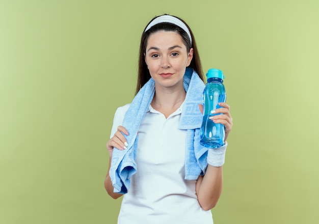 Mulher jovem fitness com fita para a cabeça e toalha em volta do pescoço segurando uma garrafa de água e sorrindo confiante em pé sobre a parede de luz