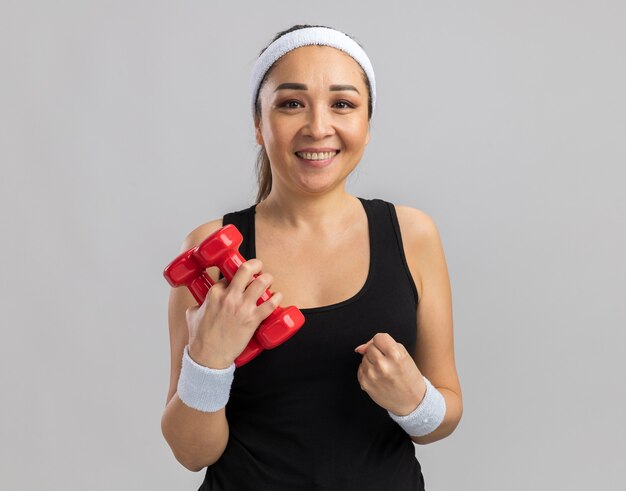 Mulher jovem fitness com fita para a cabeça segurando halteres, fazendo exercícios com os punhos cerrados, feliz e animada em pé sobre uma parede branca