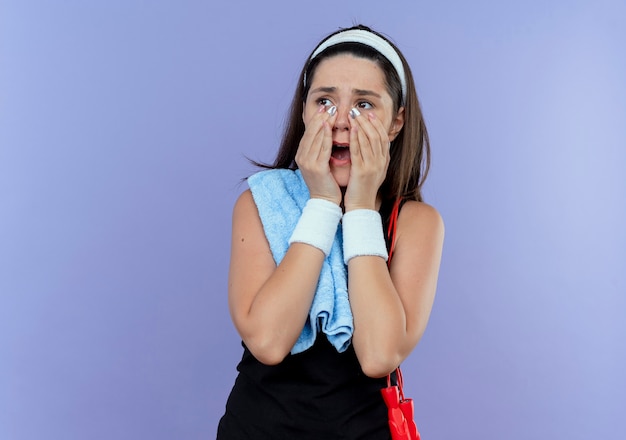 Mulher jovem fitness com uma bandana e uma toalha no ombro olhando para o lado chocada com a boca escancarada de pé sobre a parede azul