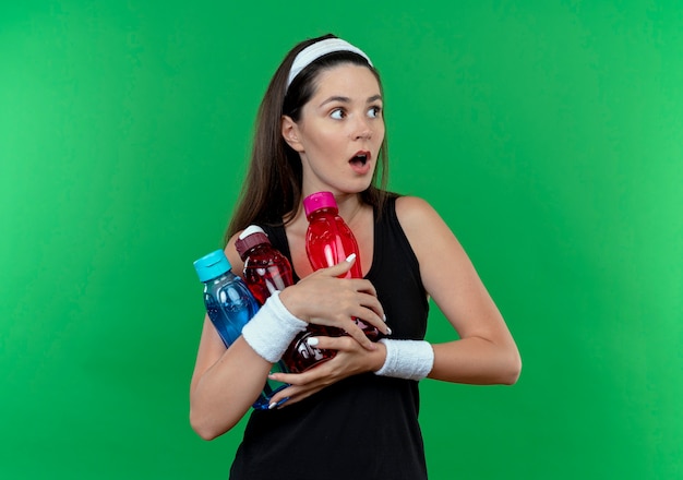 Mulher jovem fitness com uma bandana segurando garrafas de água olhando para o lado com expressão de medo em pé sobre um fundo verde