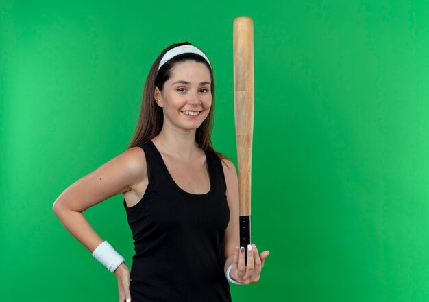 Mulher jovem fitness com uma bandana segurando um taco de beisebol, olhando para a câmera, sorrindo com uma cara feliz em pé sobre um fundo verde