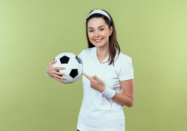 Mulher jovem fitness com uma bandana segurando uma bola de futebol apontando com o dedo para ela e sorrindo alegremente em pé sobre a parede de luz