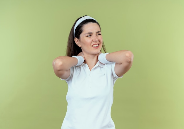 Mulher jovem fitness com uma bandana tocando seu pescoço, olhando para o lado, sentindo desconforto em pé sobre um fundo claro