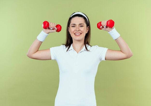 Mulher jovem fitness com uma faixa na cabeça e malhando com halteres esticados em pé sobre a parede de luz